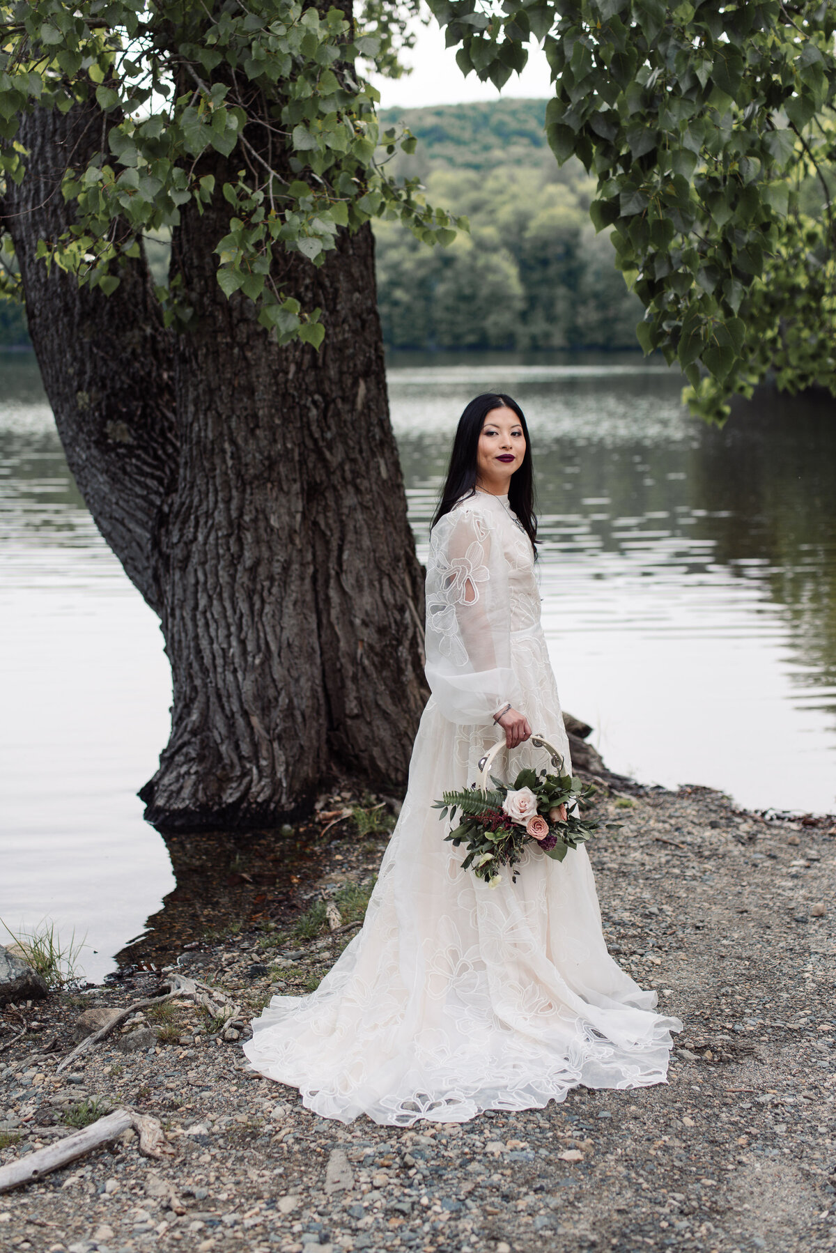 Bride wearing bespoke Daarlana wedding gown at a lake in VT. Holding tambourine bouquet by haven Floral company.