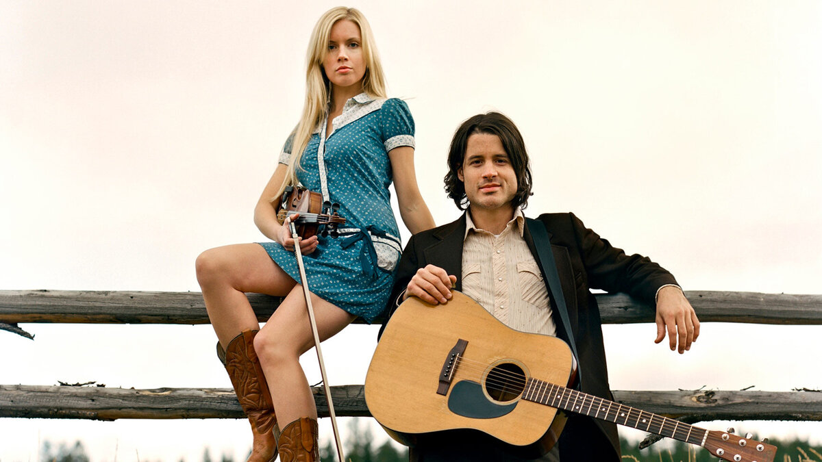 Country Music Duo Portrait Dustin Bentall Kendel Carson Dustin standing and leaning on wood fence with guitar Kendel sitting on wood fence with fiddle in her lap