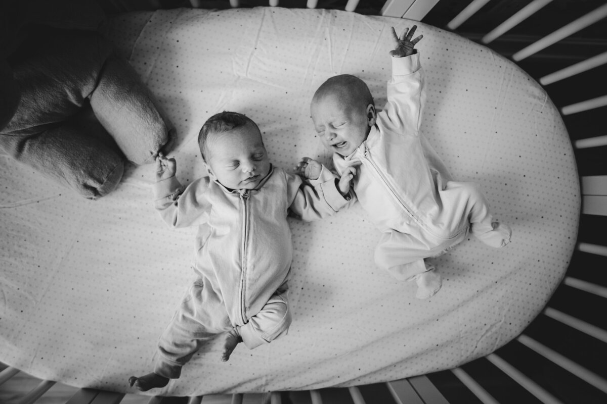 Overhead photo of two newborn twins fussing in a crib. This image is black and white.