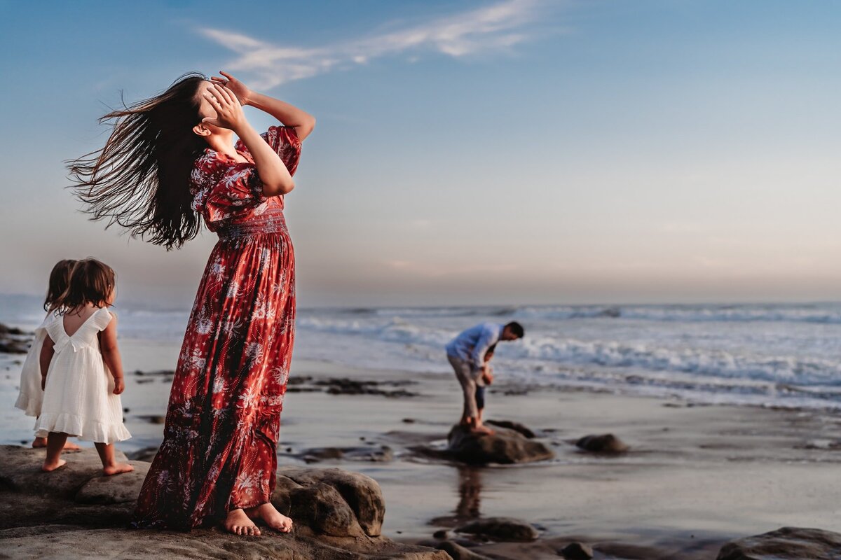 A woman stands on a rock on a Del Mar beach, whipping her dark hair around, while her family plays behind her. This is during a lifestyle beach photography session by San Diego photographer Love Michelle Photography
