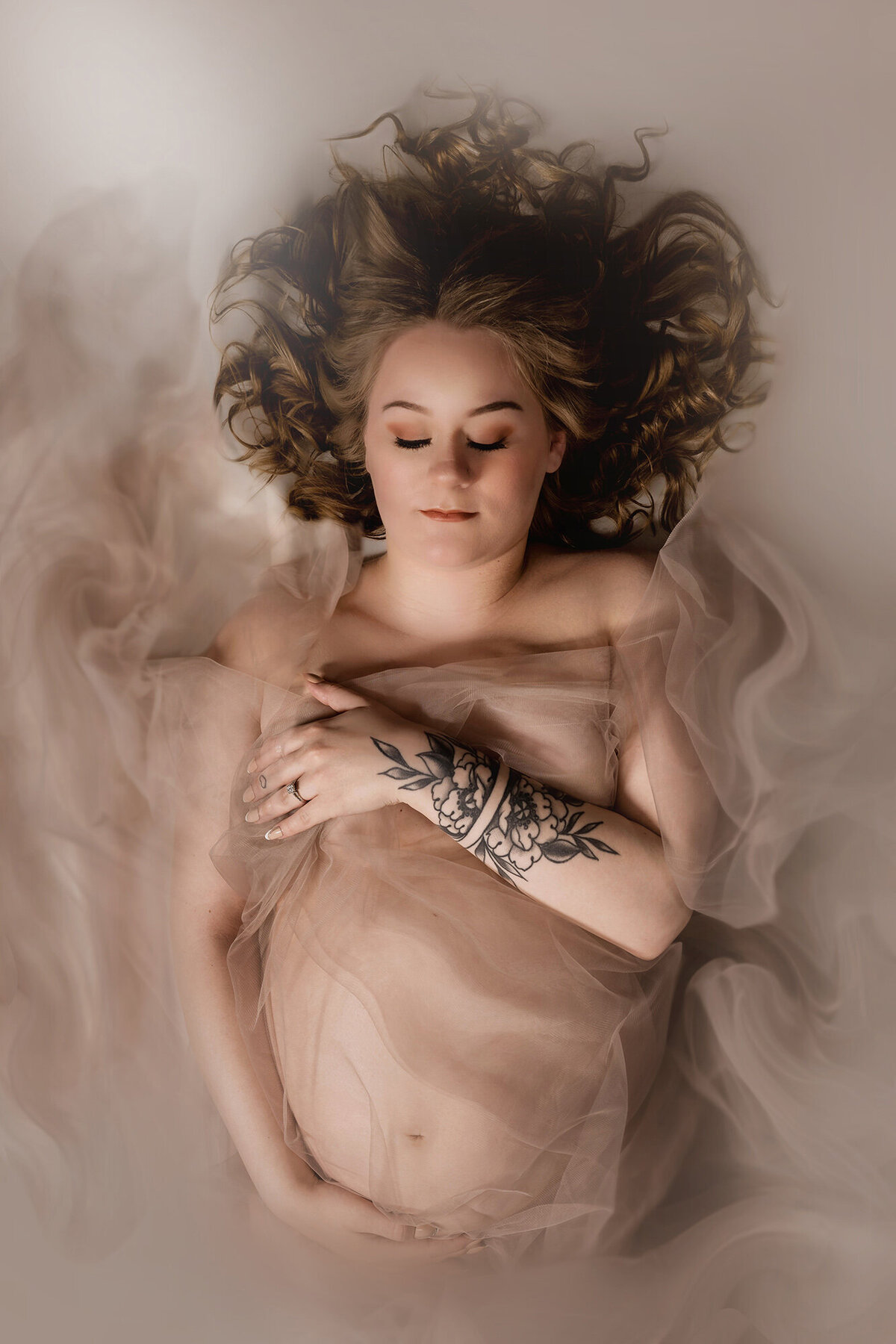 A pregnant woman lays in a studio covering herself with her hands and a sheer fabric