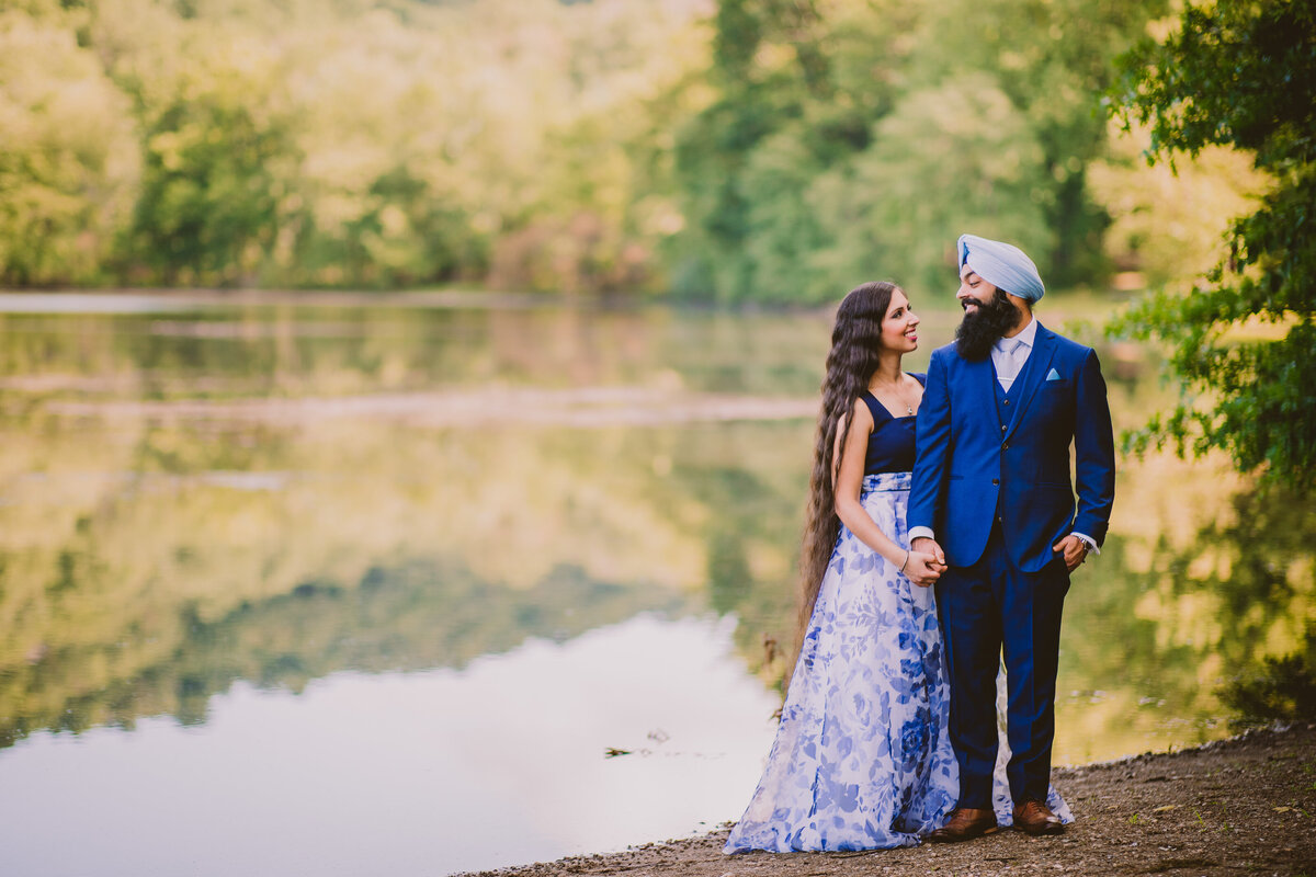 An engaged sikh couple in blue outfits smile at each other near the lake at ramapo valley reservation in nj