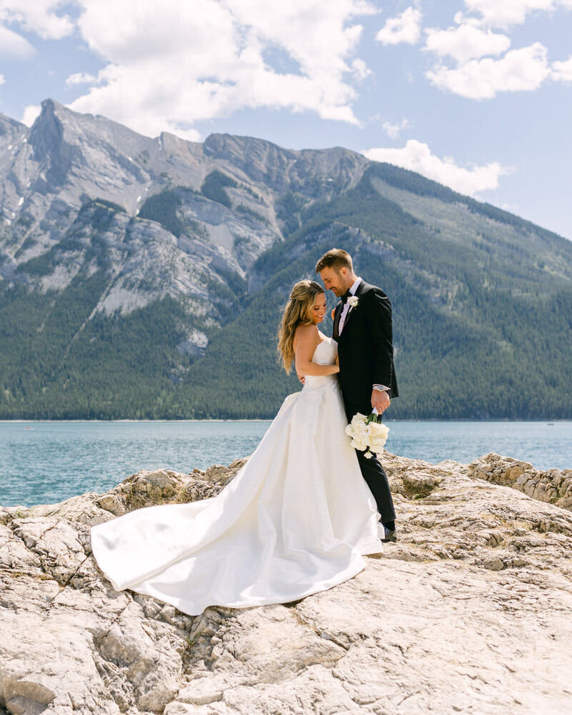 This breathtaking photograph captures the bride and groom holding each other on a rock at the shoreline of Lake Minnewanka in Banff National Park. The stunning blue waters and rugged mountains create a majestic backdrop, adding luxury and romance to their special day.