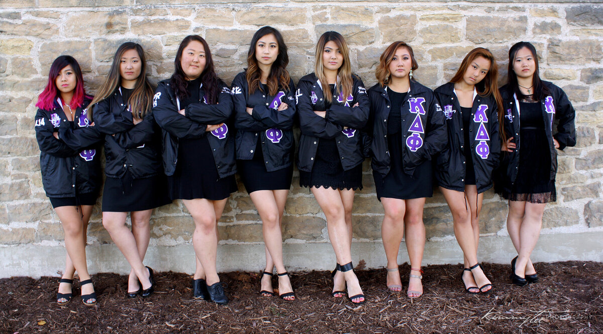 Eight sorority girls in line jackets standing against brick wall