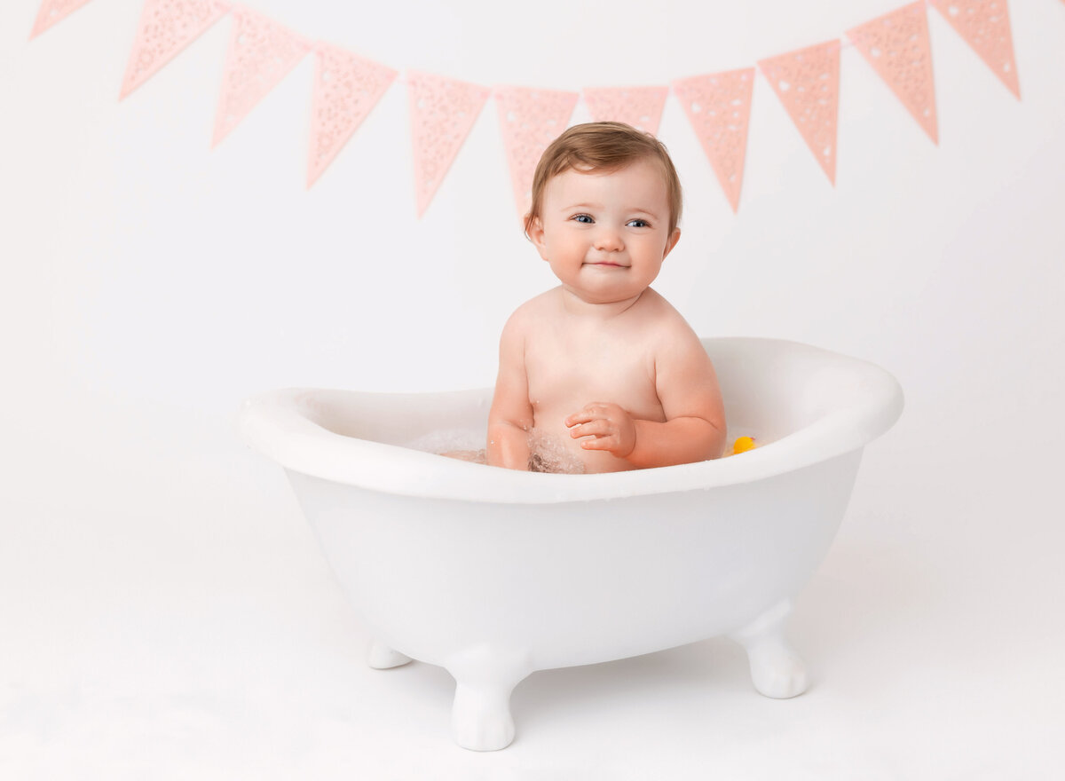 Baby girl sits in bathtub smiling at the camera after a messy cake smash photoshoot.