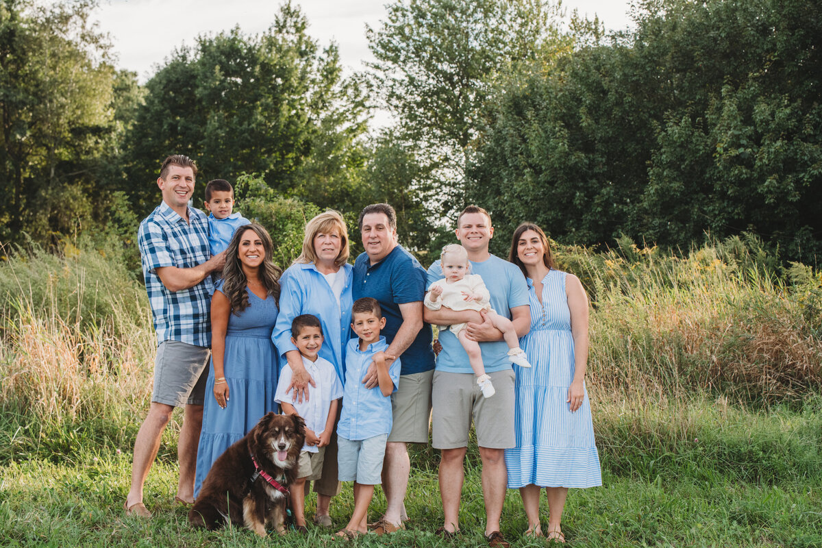 A multigenerational family dressed in coordinated blue outfits poses together in a lush green field with their beloved dog during an outdoor family photography session in Connecticut. Captured by Nicole Mele Photography, highlighting love, togetherness, and lasting memories.