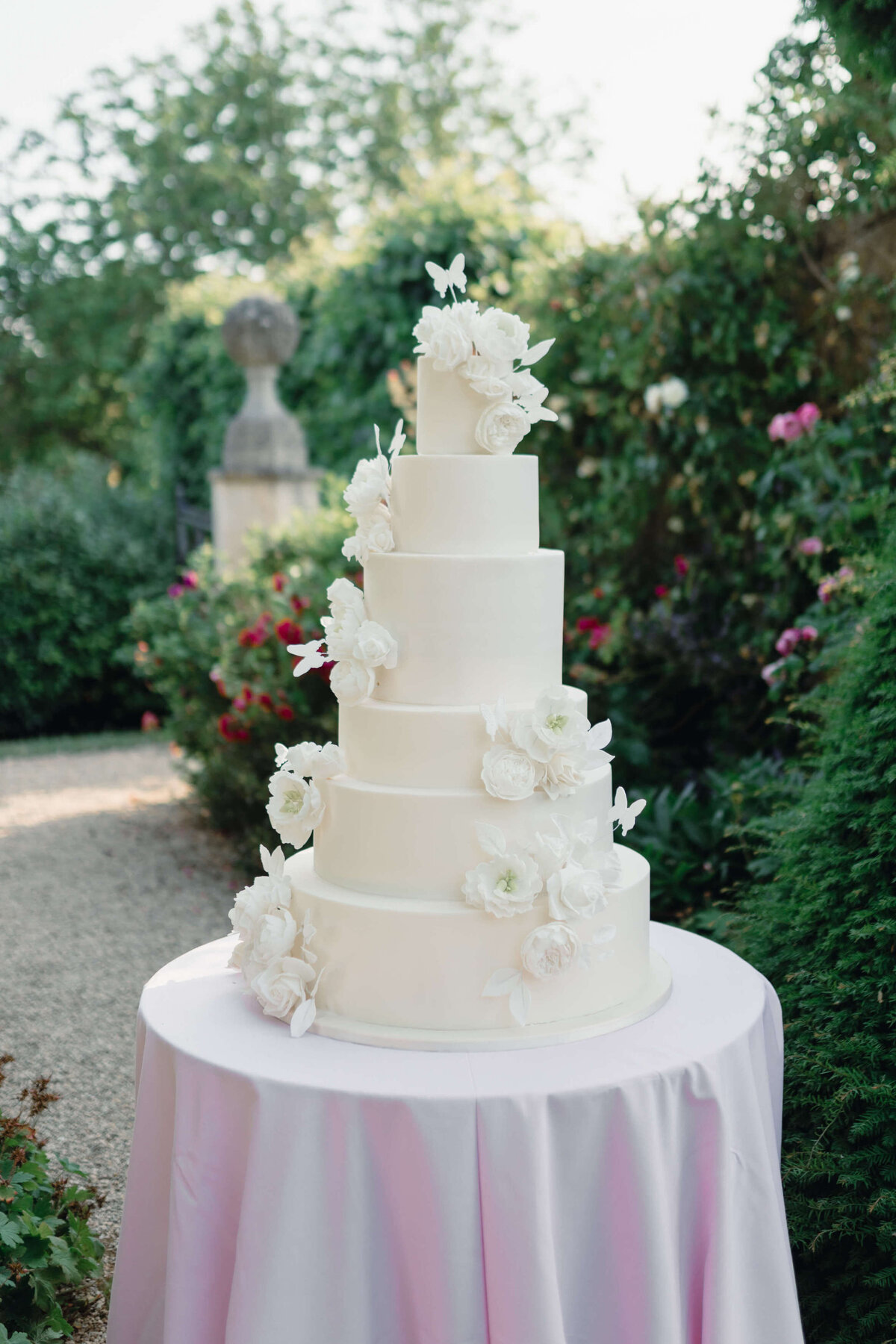 romantic six tier white wedding cake decorated with sugar flowers and butterflies sat on pink table linen in the gardens of euridge manor