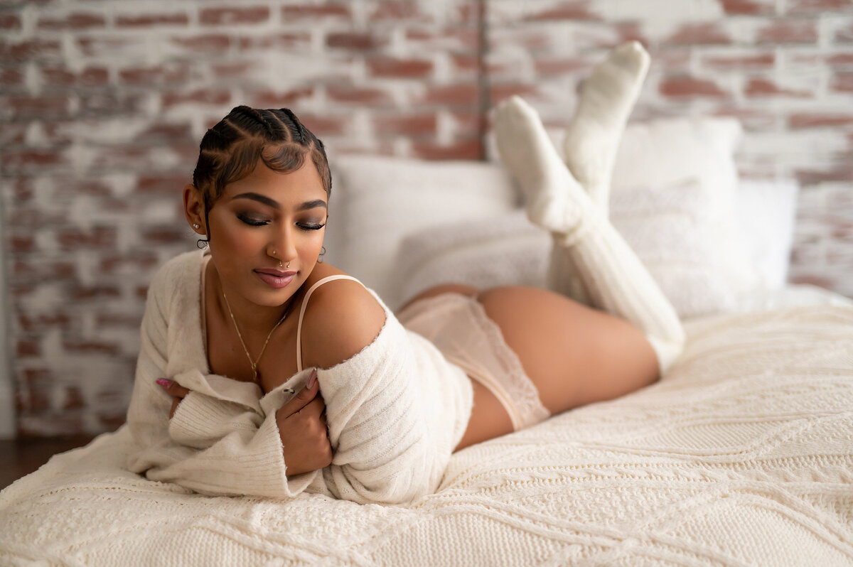 A woman lays on a bed in our Waukesha  studio wearing a knit sweater and knee-highs for a cozy boudoir portrait session.