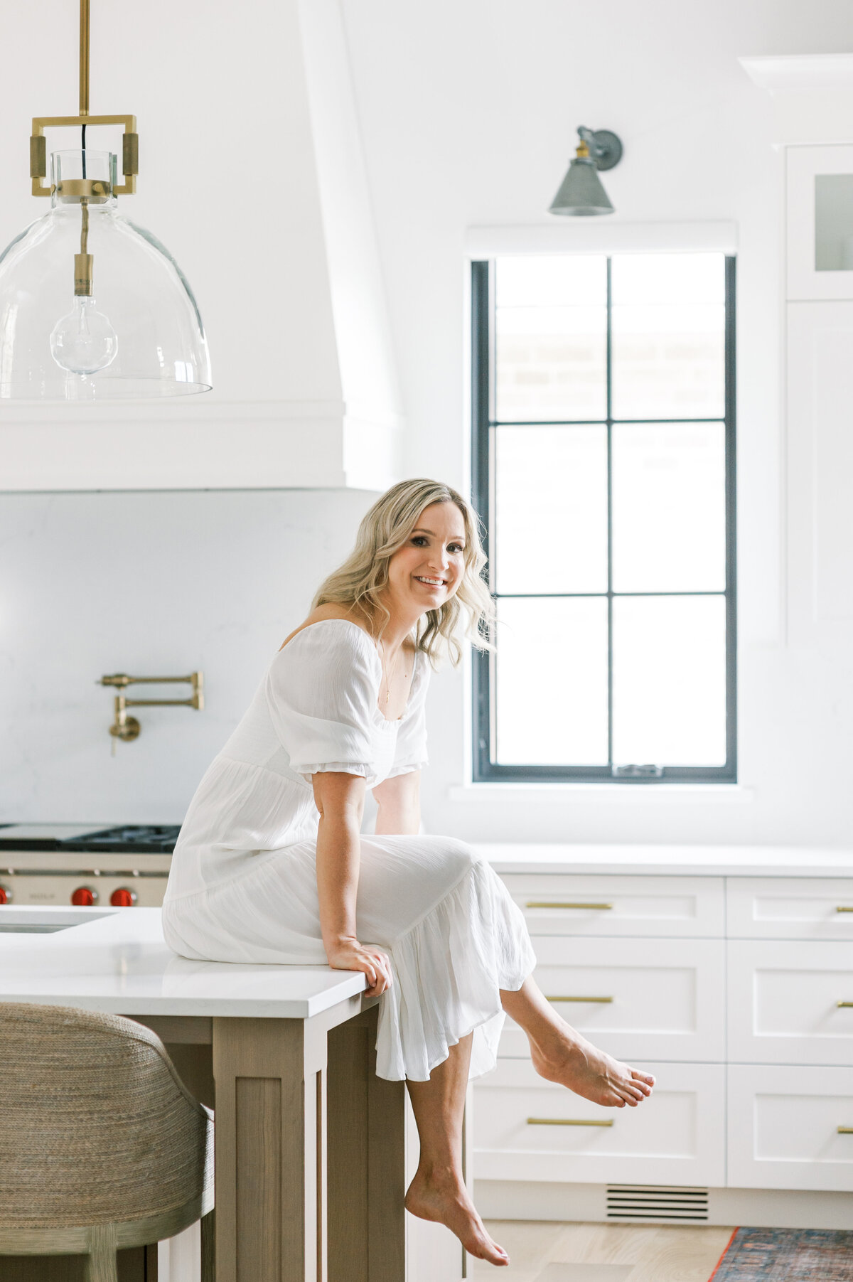 Premiere Windsor, Ontario Interior Designer sits casually atop of her countertop with her legs crossed. She is looking over her shoulder and smiling at the camera. Her all white kitchen and black framed windows are behind her.