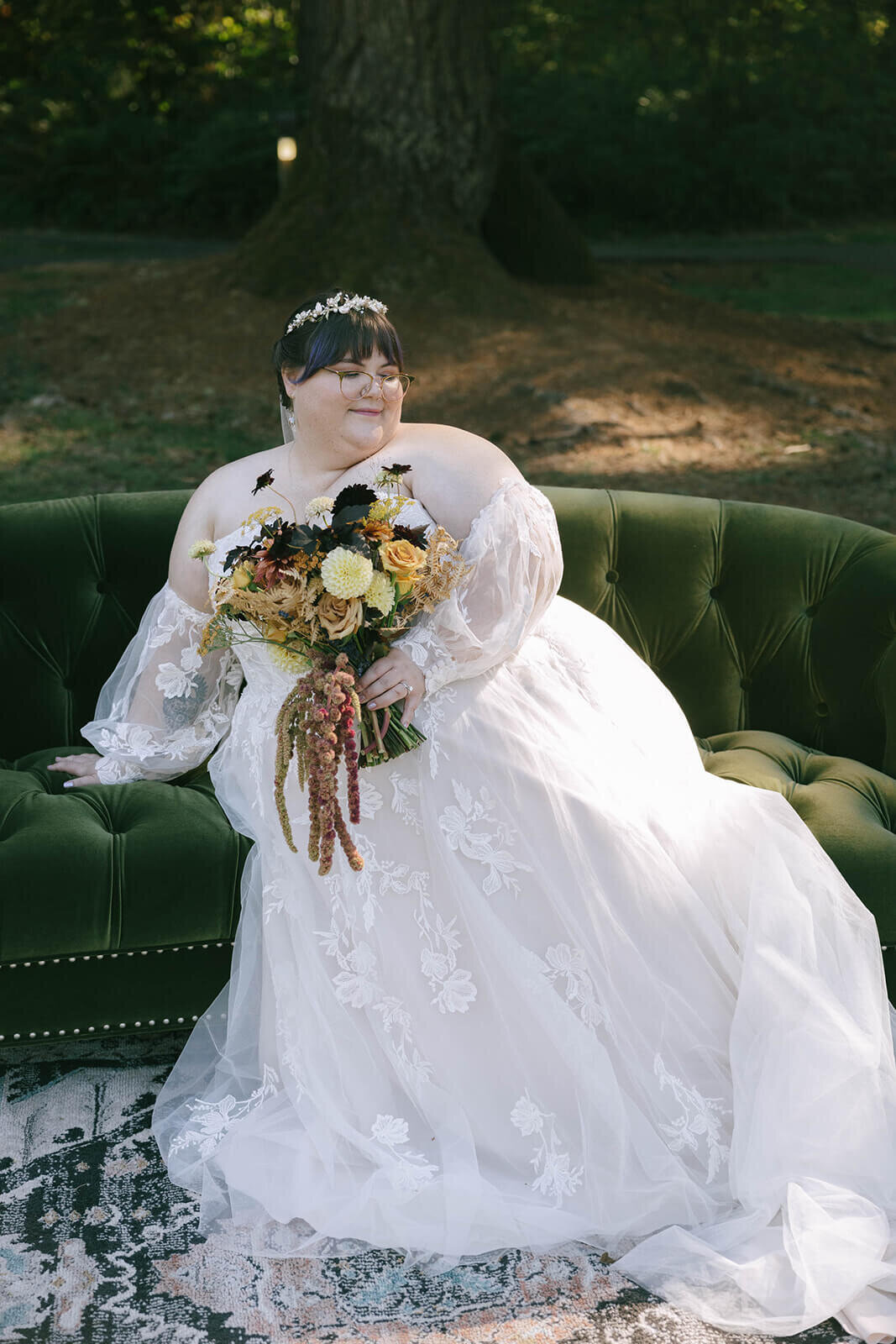 A bride leans back while seated on a green couch and holds her flower bouquet.