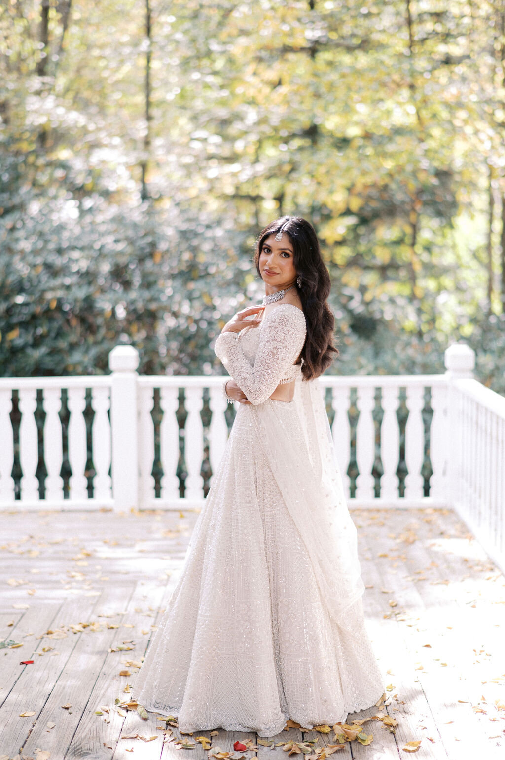 Full-length bridal portrait outdoors on a porch with fall leaves and trees behind her.