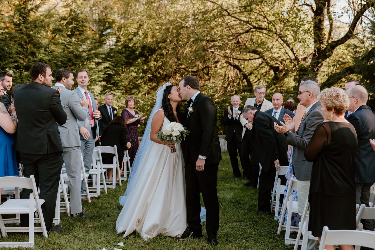 A bride and groom share a kiss outside the Stagecoach Inn, standing beside a bush of blooming hydrangeas. The bride wears a classic white gown with a deep V-back and a long train, while the groom is dressed in a black tuxedo. The couple is framed by the white brick exterior of the inn and the lush greenery, creating a romantic and timeless scene.