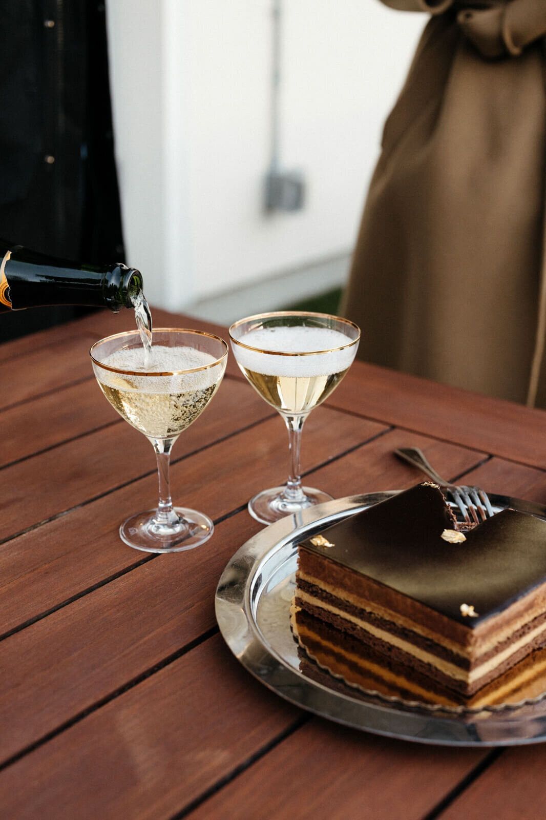 Two wine glasses and a slice of chocolate cake are on a table. The bottle of champagne is being poured over one wine glass.