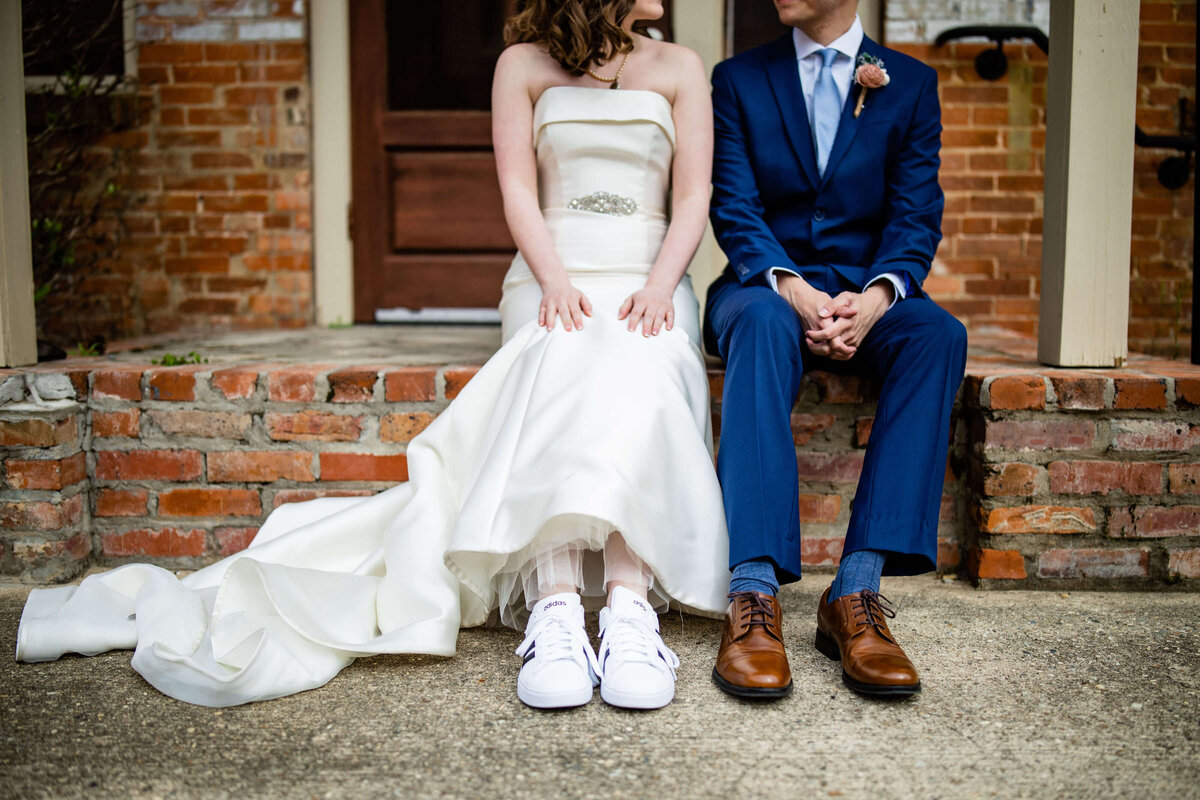 A bride in a white dress and sneakers sits beside a groom in a blue suit on a brick ledge outside a brick building. They are holding hands, smiling, and looking at each other.