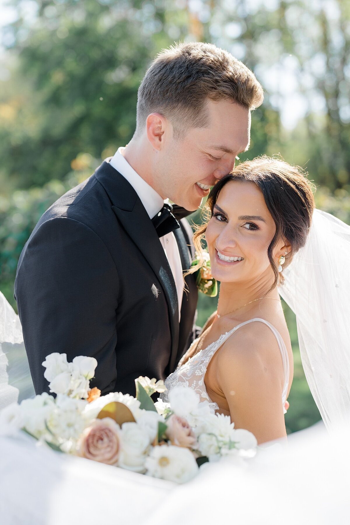 bridal portrait with groom holding his bride and smiling as he leans his face into her head while the brides veil flows in the wind and she smiles over her shoulder