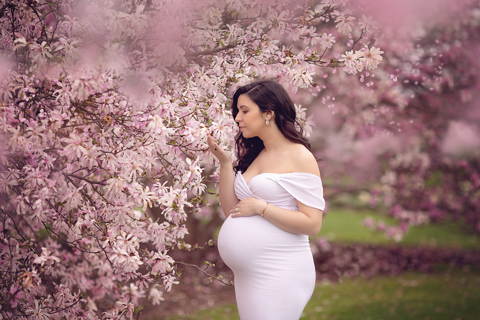 A pregnant woman in a pink off the shoulder maternity gown smells some pink blossoms in a park posed by a New Jersey Maternity Photographer