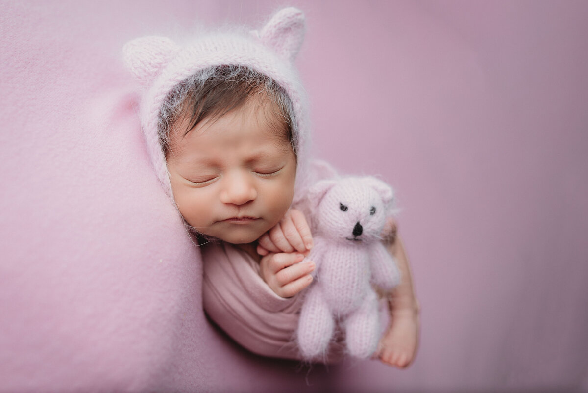 2 week old newborn girl sleeping and holding stuffed bunny and wearing a purple swaddle and white bonnet with ears on it