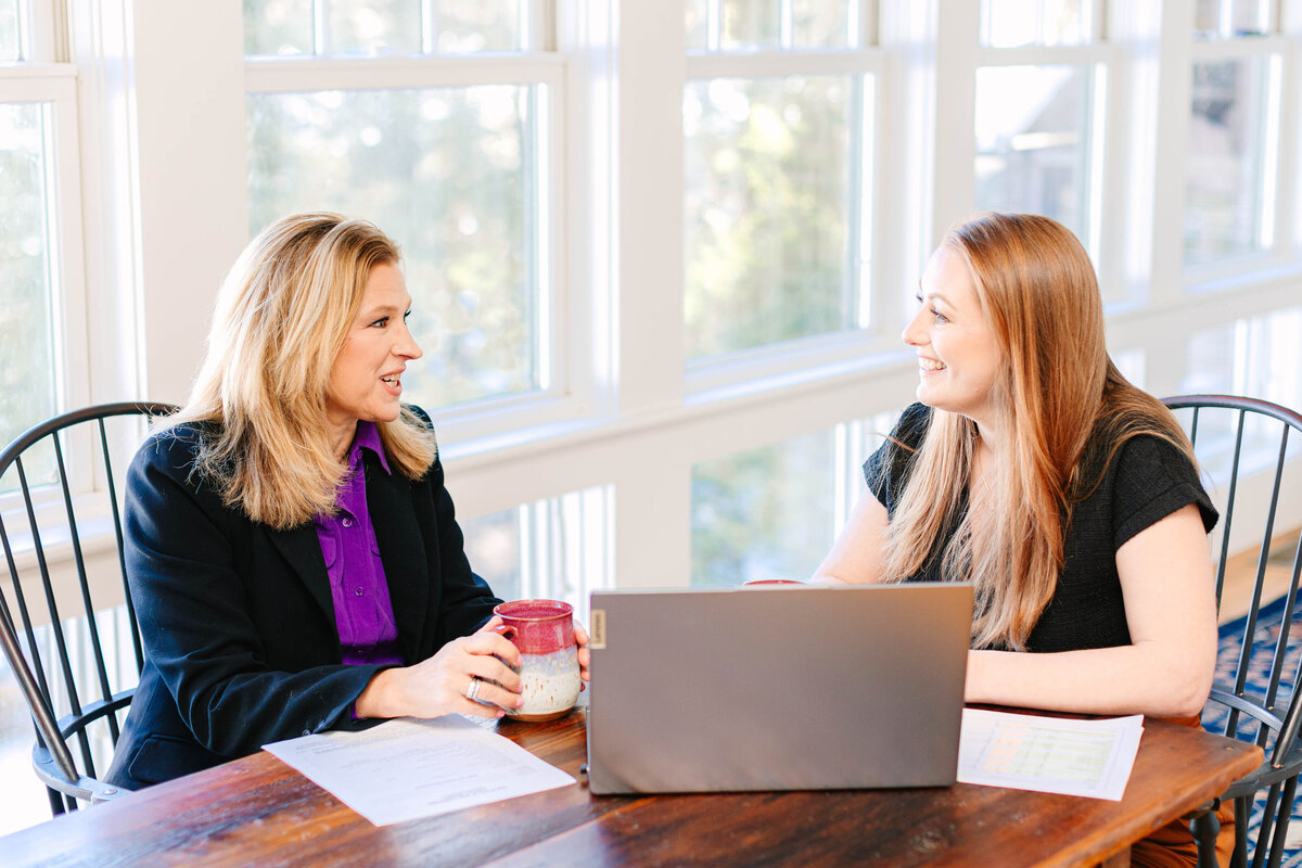Two professional women sit at a wooden table in a bright office space, engaged in conversation. One holds a coffee mug, while the other smiles with an open laptop in front of her. Papers and documents are spread out on the table.