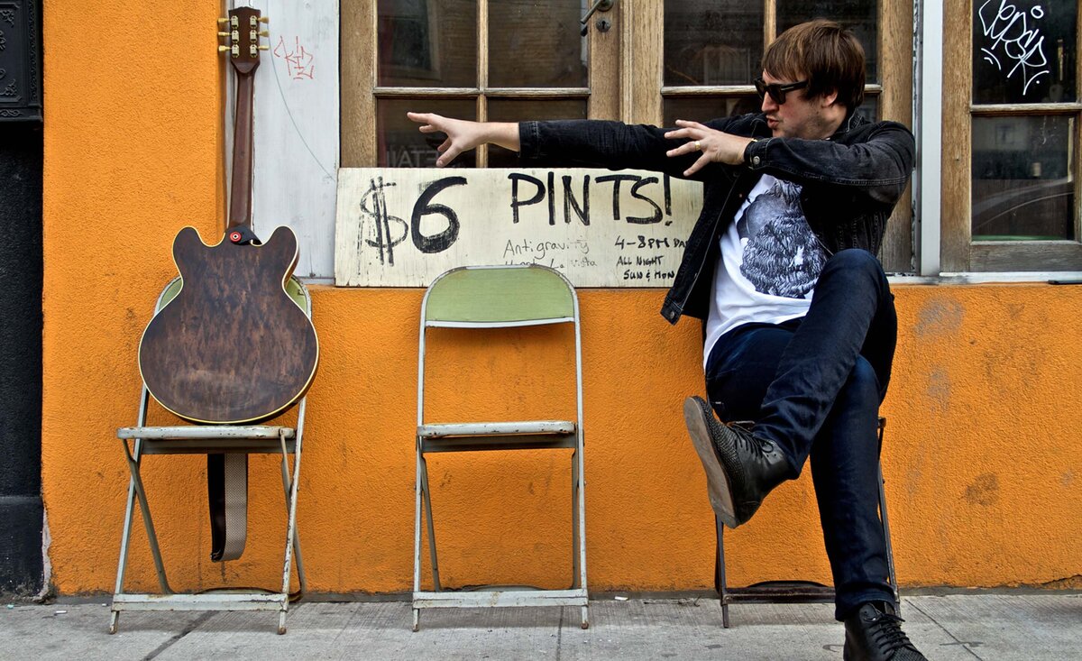 Male musician portrait Mikey Manville wearing jeans white t shirt jean jacket sitting stretching arms toward dark wood electric guitar orange pub wall behind