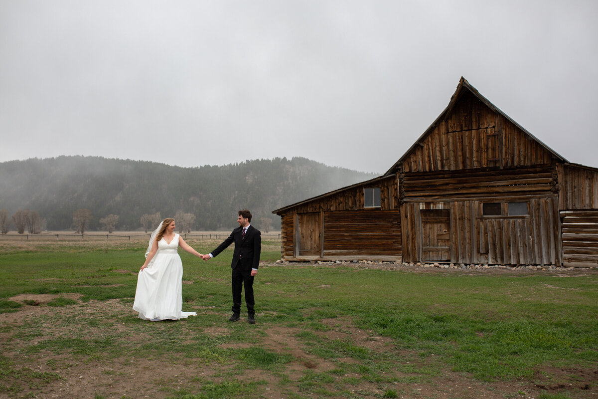 A bride and groom hold hands and stand in front of an old wooden barn in Wyoming.