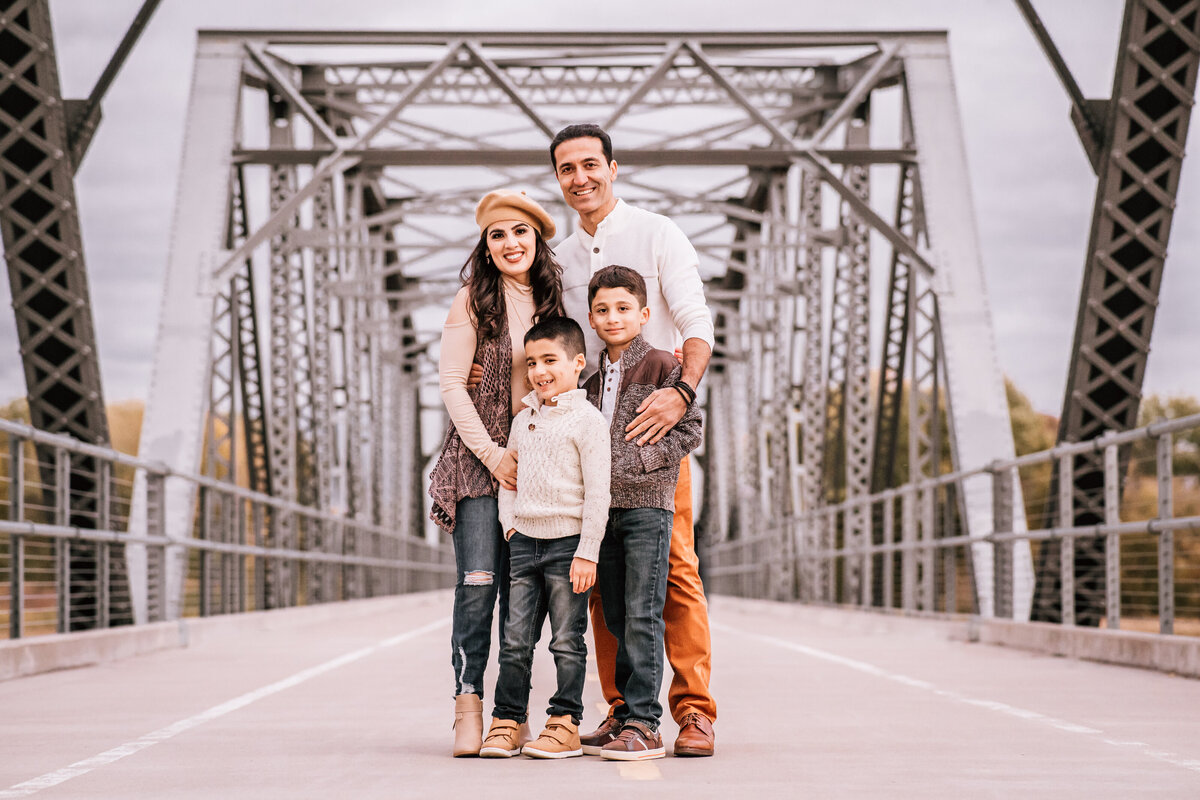 A family of four stands together in the middle of a metal bridge. The parents stand behind their two young sons, all dressed in warm, casual clothing, with the bridge's framework creating a striking background.