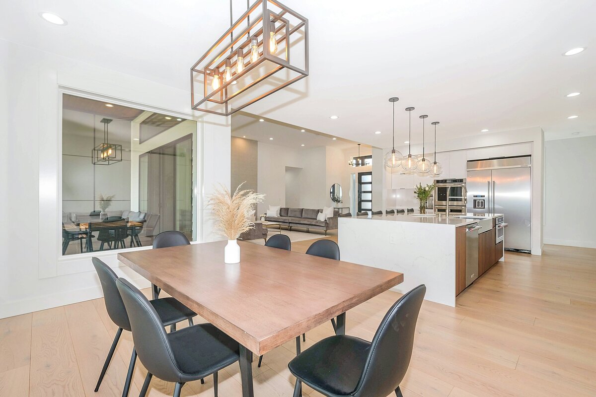 A modern, open-concept home interior featuring a dining area in the foreground and a living room and kitchen visible beyond. The dining area has a rectangular wooden table with six black chairs, illuminated by a large, rectangular industrial-style chandelier. Light wood floors extend throughout the space. In the background, a spacious living room features a dark gray sectional sofa and a large, brightly lit kitchen with a white island, pendant lights, and built-in appliances. The overall aesthetic is clean, contemporary, and airy.