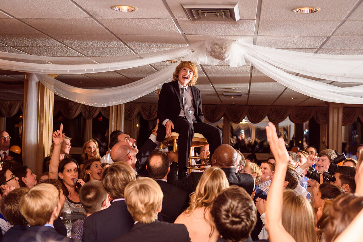 A boy in a suit is lifted up on a chair in the middle of a busy dancefloor