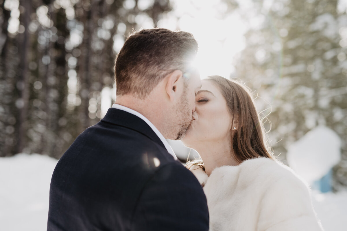 grand teton wedding with bride and groom kissing as the sun shines over head through the trees in the woods for their winter elopement
