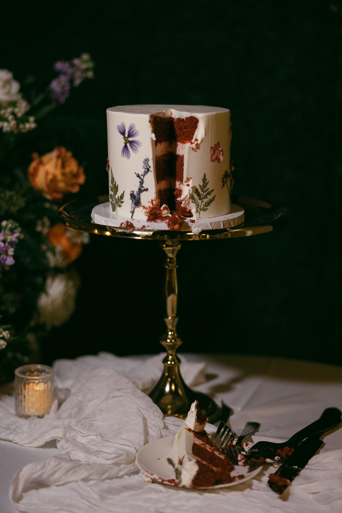 A small white cake with florals on the outside with one slice cut out of it stands on a gold platter atop a table with florals in the background