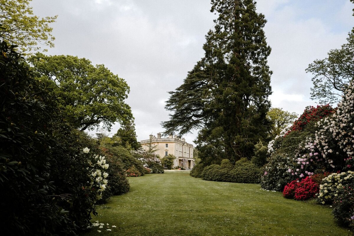 An idyllic English country garden in soft whites and pinks, framed by a towering tree and the timeless elegance of a blonde stone Georgian manor.
