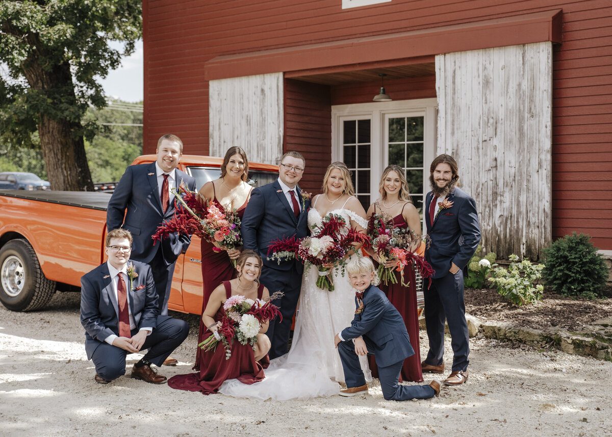 A joyous wedding party posing for a photo in front of a rustic red barn, with the bride and groom surrounded by their bridesmaids and groomsmen, each holding elegant bouquets, and a classic orange pickup truck in the background adding a touch of vintage charm to the countryside celebration taken by Jen Jarmuzek Phpotography a minneapolis wedding photographer