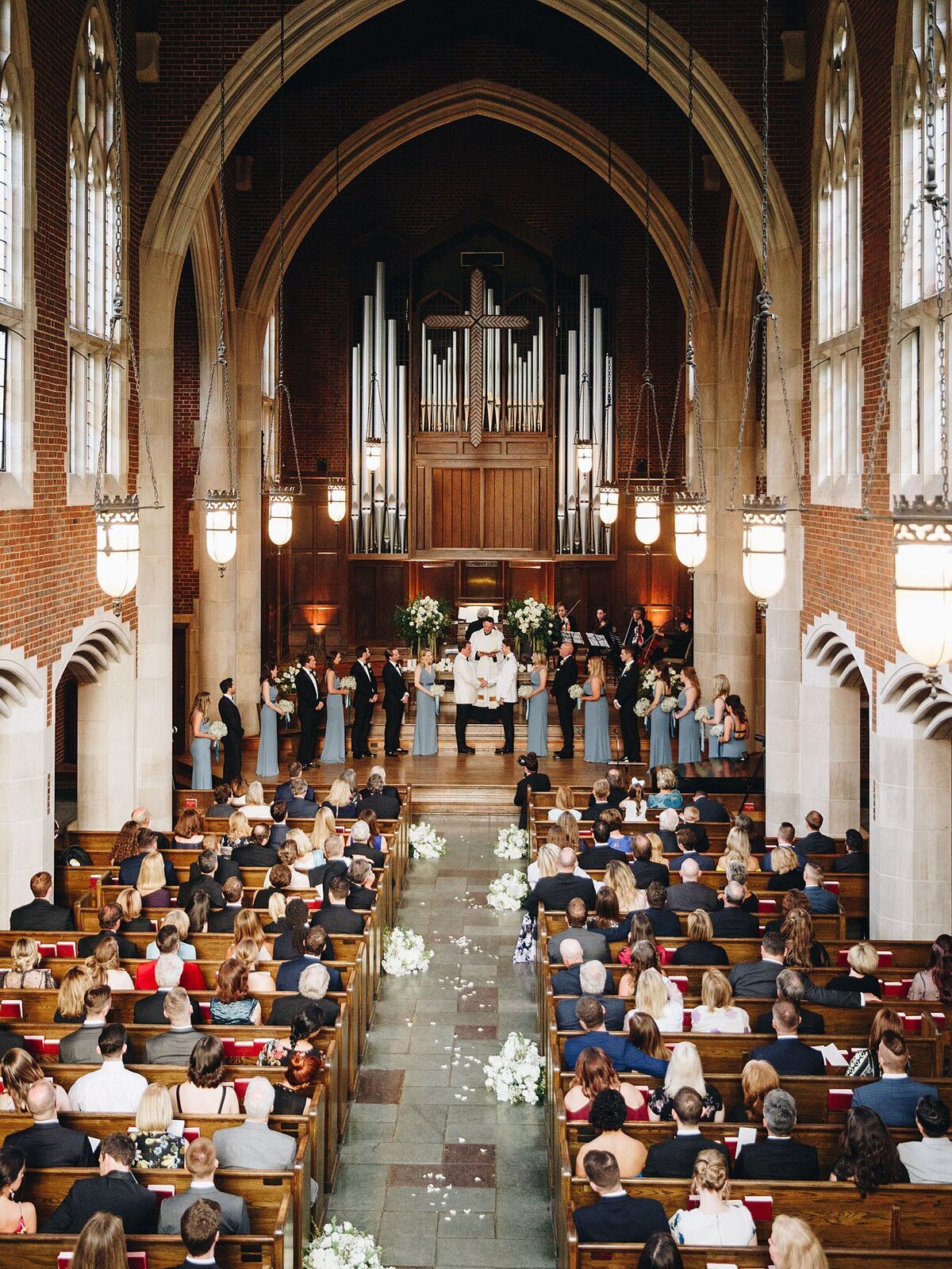 Wedding ceremony for two grooms wearing white tuxedo jackets and black pants at Scarritt Bennet. The stone church aisle is lined with large round white floral arrangements. The church pews are filled with wedding guests and there is a large pipe organ behind the altar. On either side of the grooms is the wedding party dressed in dusty blue dresses and black tuxedos.