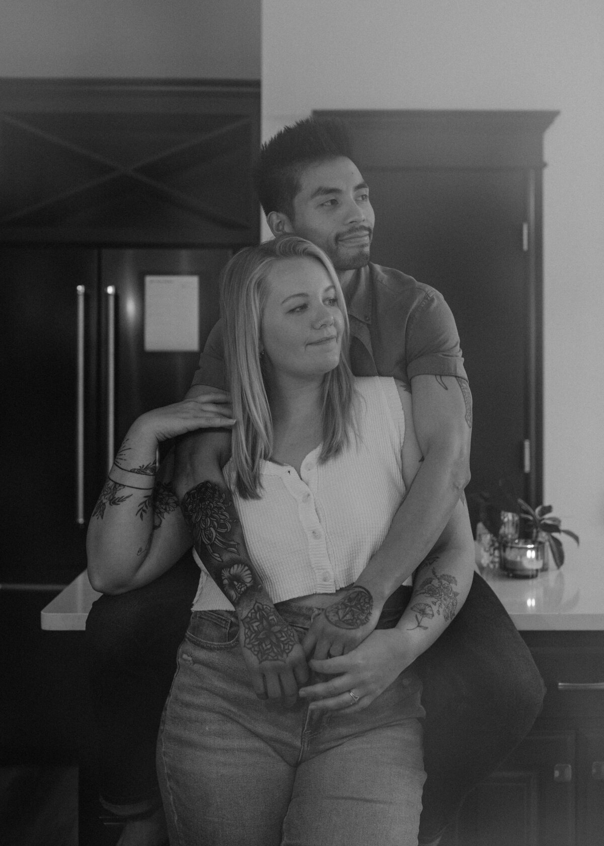 Black and white image of a couple embracing in a kitchen, with the person behind resting their chin on the other's shoulder
