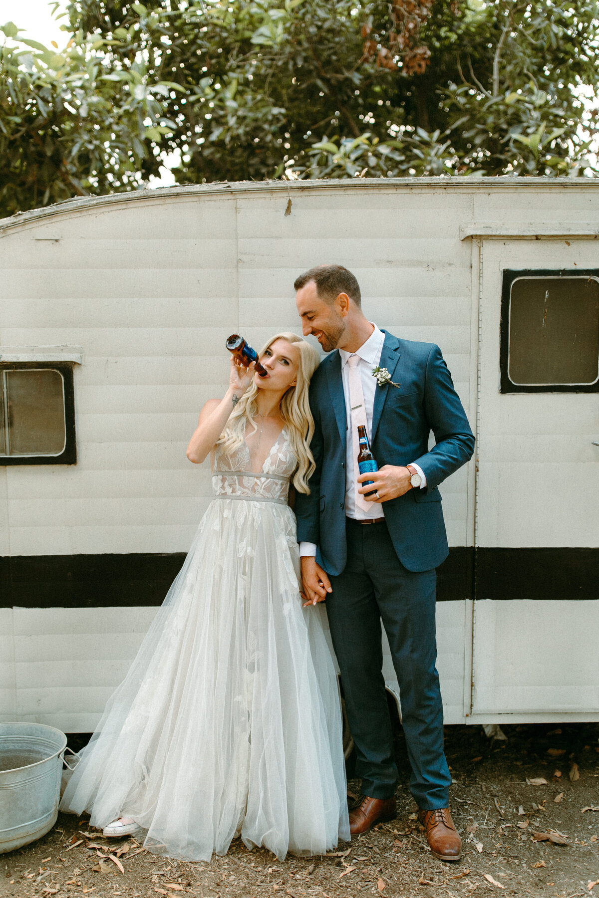 A bride and groom posing and drinking beers together holding hands.