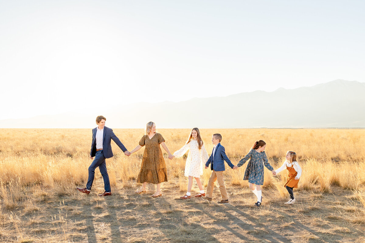 A family of six walks hand-in-hand through a golden field at sunset in Eagle Mountain, Utah, with mountains in the background.