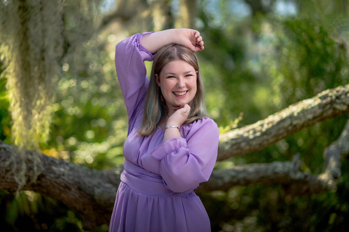 A high school grad in a purple dress stands in a forest with a hand over her head