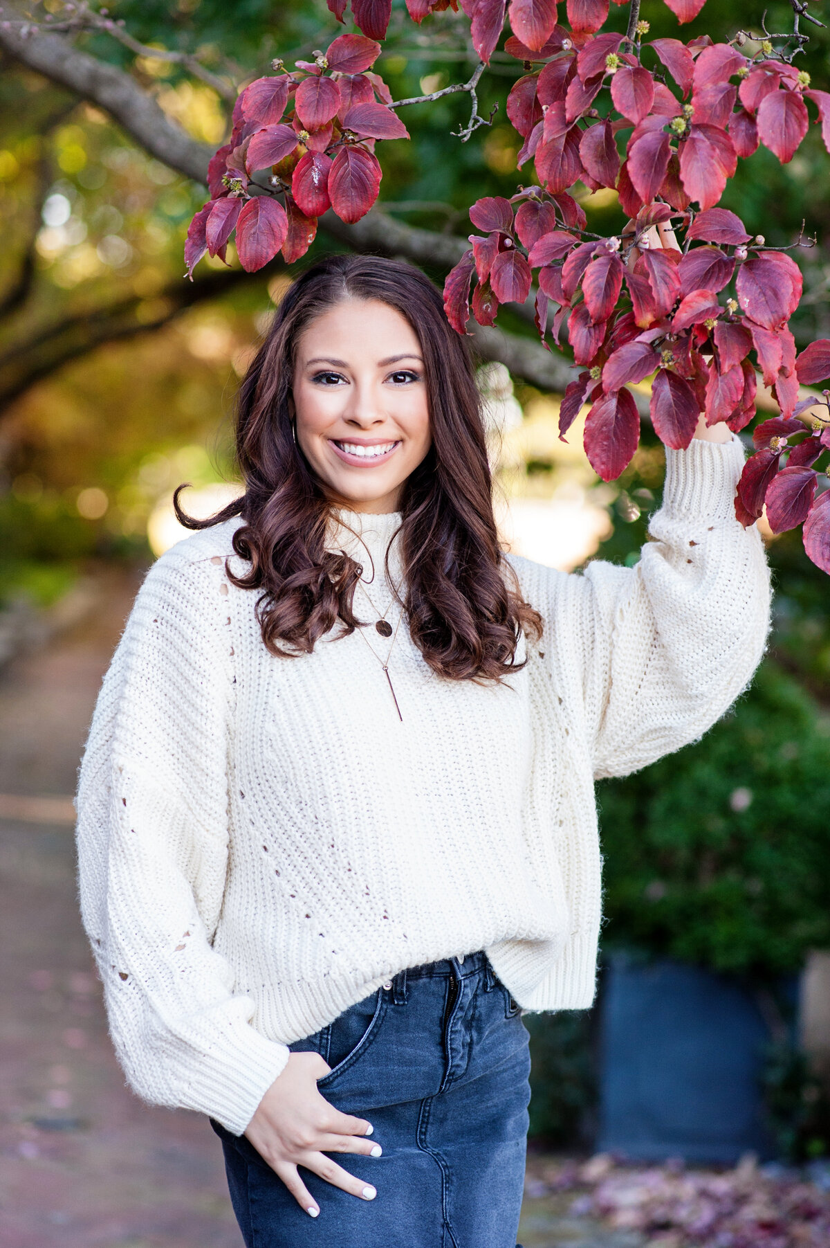 Mechanicsville high school senior girl poses during fall with red leaves. She is wearing a denim skirt and beige sweater in Libby Hill Park.