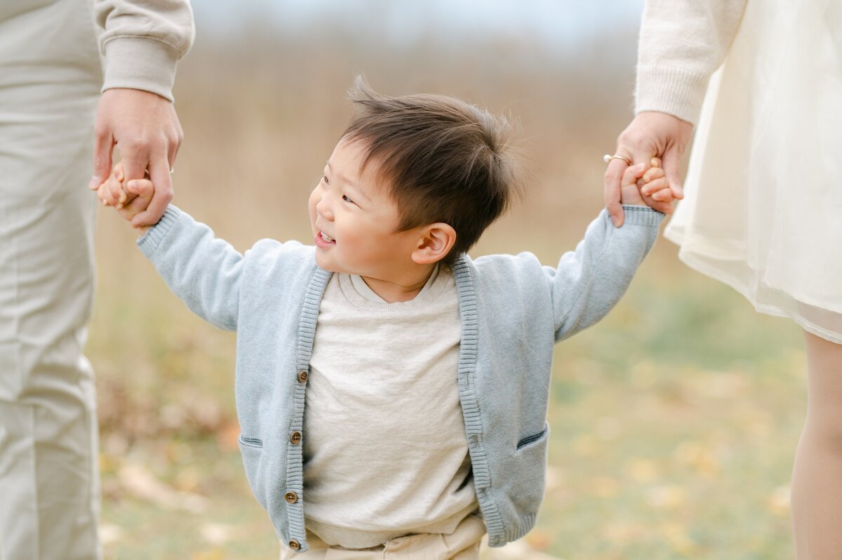 Close up photograph of a little boy who is holding hands with both of his parents and smiling to the side