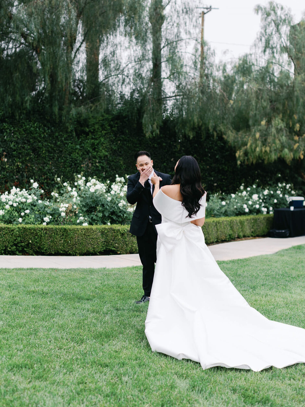 Bride in a flowing white gown with a large bow stands facing groom on a lush green lawn. Groom is emotional, smiling.