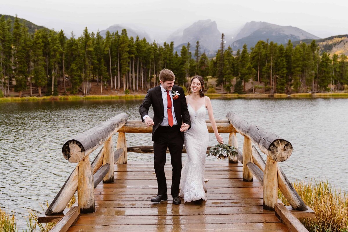 Bride and groom walking hand in hand across a wooden dock during their wedding at Sprague Lake in Rocky Mountain National Park, with pine trees and mountain peaks in the background.