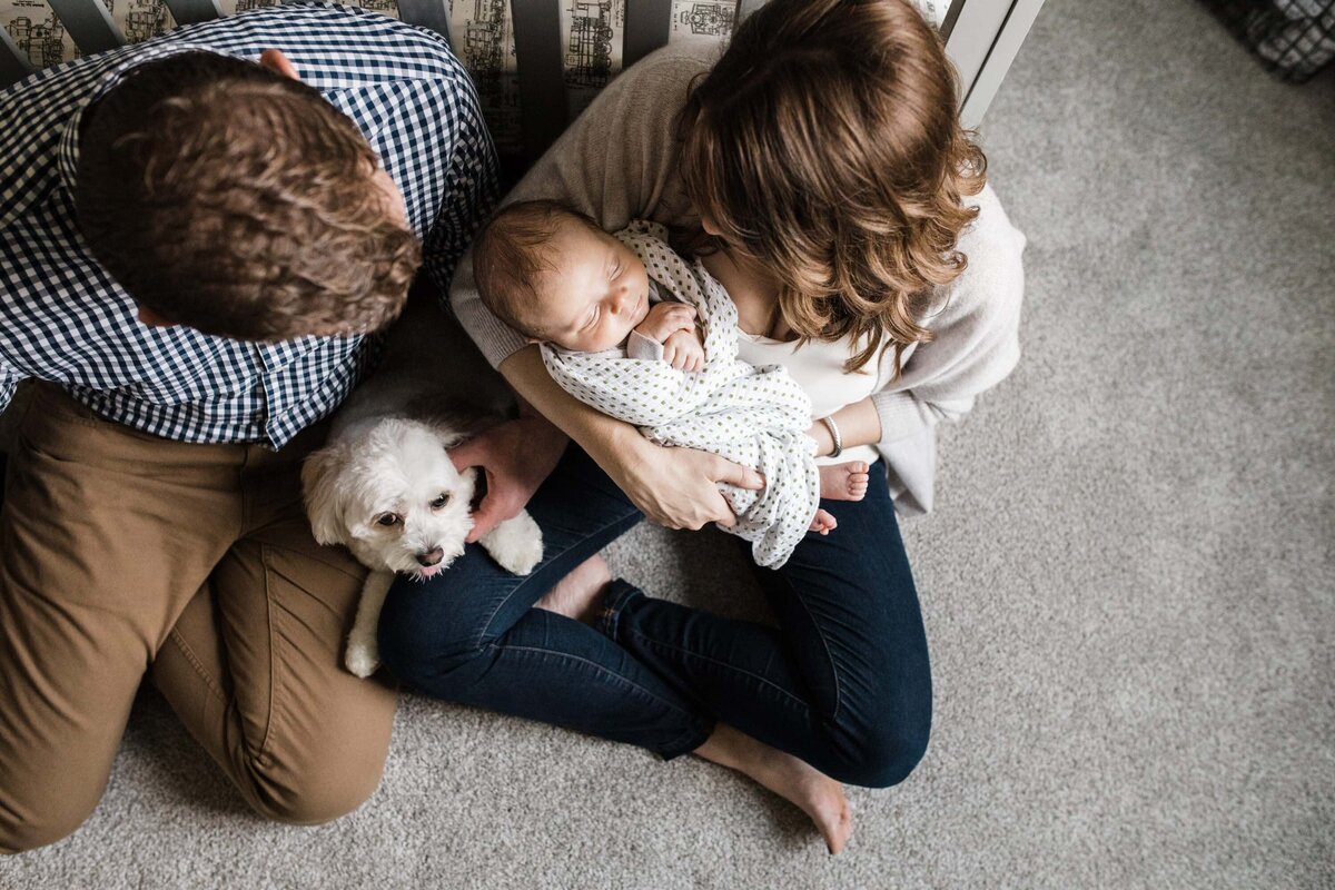 Family and pet dog sitting on the floor at home, with parents looking affectionately at a sleeping baby in the mother's arms during a newborn photography session.