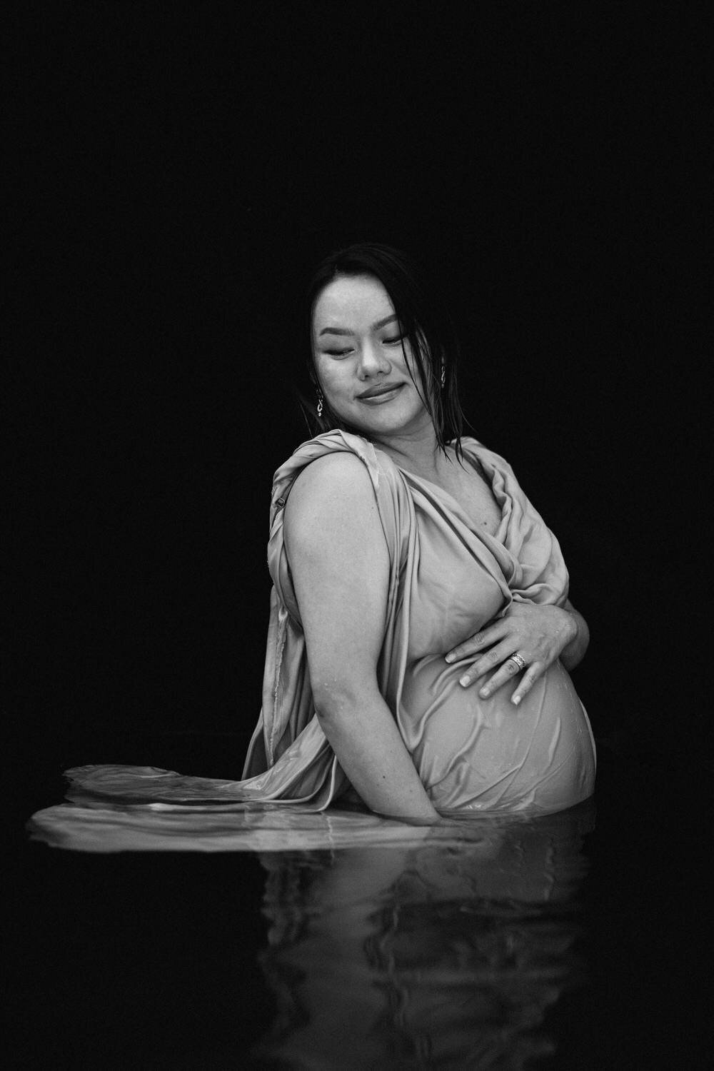 A pregnant Asian woman stands in pool with a black background and holds her bump