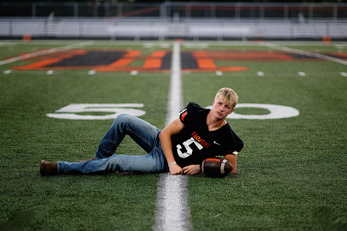 A high school senior lays across the 50 yard line in his jersey and jeans with a ball as guided by his Pella Senior Photographer