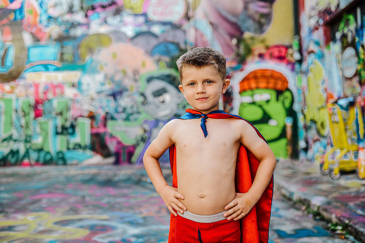 Little boy with no shirt wearing a red superhero cape with hands on hips at graffiti alley near MICA in Baltimore Maryland