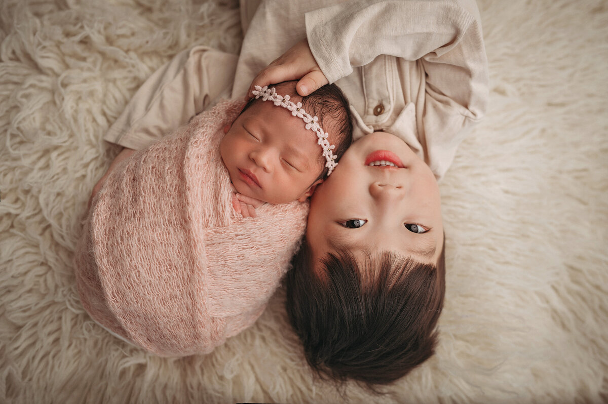 smiling new big  brother  laying down on his back upside down while holding his newborn baby sister who is wrapped in a pink swaddle and wearing a floral headband facing right side up on a white rug