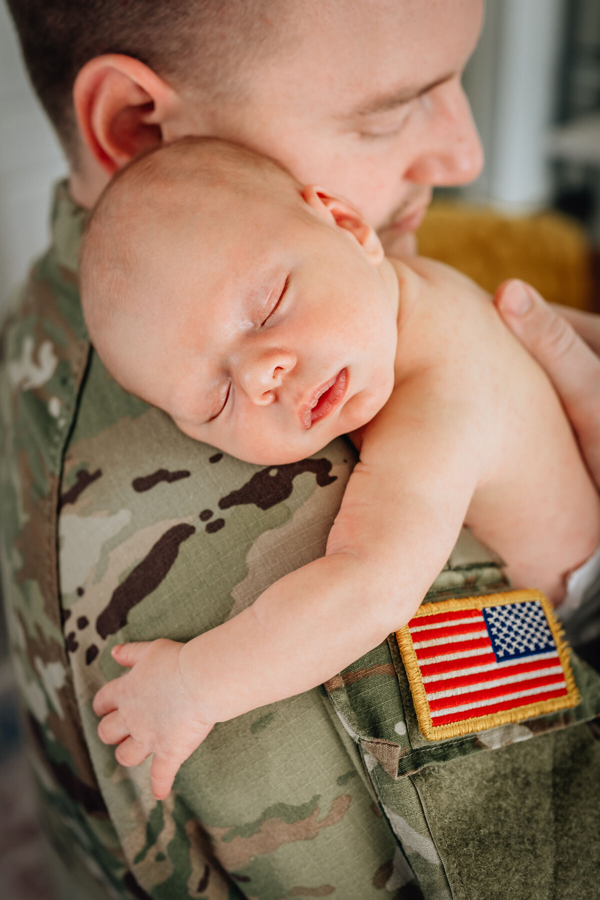A close-up of a newborn sleeping peacefully on their father's chest, who is dressed in a military uniform with an American flag patch on the shoulder.