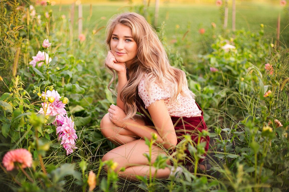 A-young-woman-with-long-blonde-hair-wearing-a-floral-top-and-maroon-skirt-crouching-among-blooming-flowers-in-a-lush-green-garden-at-golden-hour-during-her-indianapolis-senior-portrait-session-senior-photography-in-Indianapolis