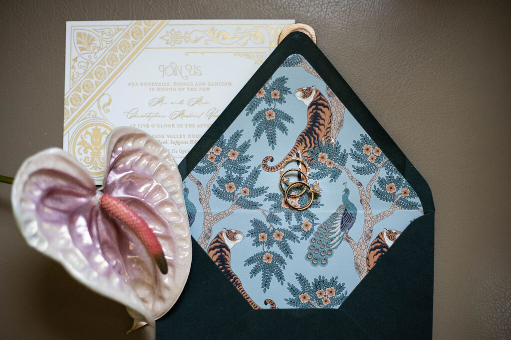 A beautifully styled wedding invitation suite featuring an intricate floral design, a deep navy envelope, and a delicate pink anthurium.
