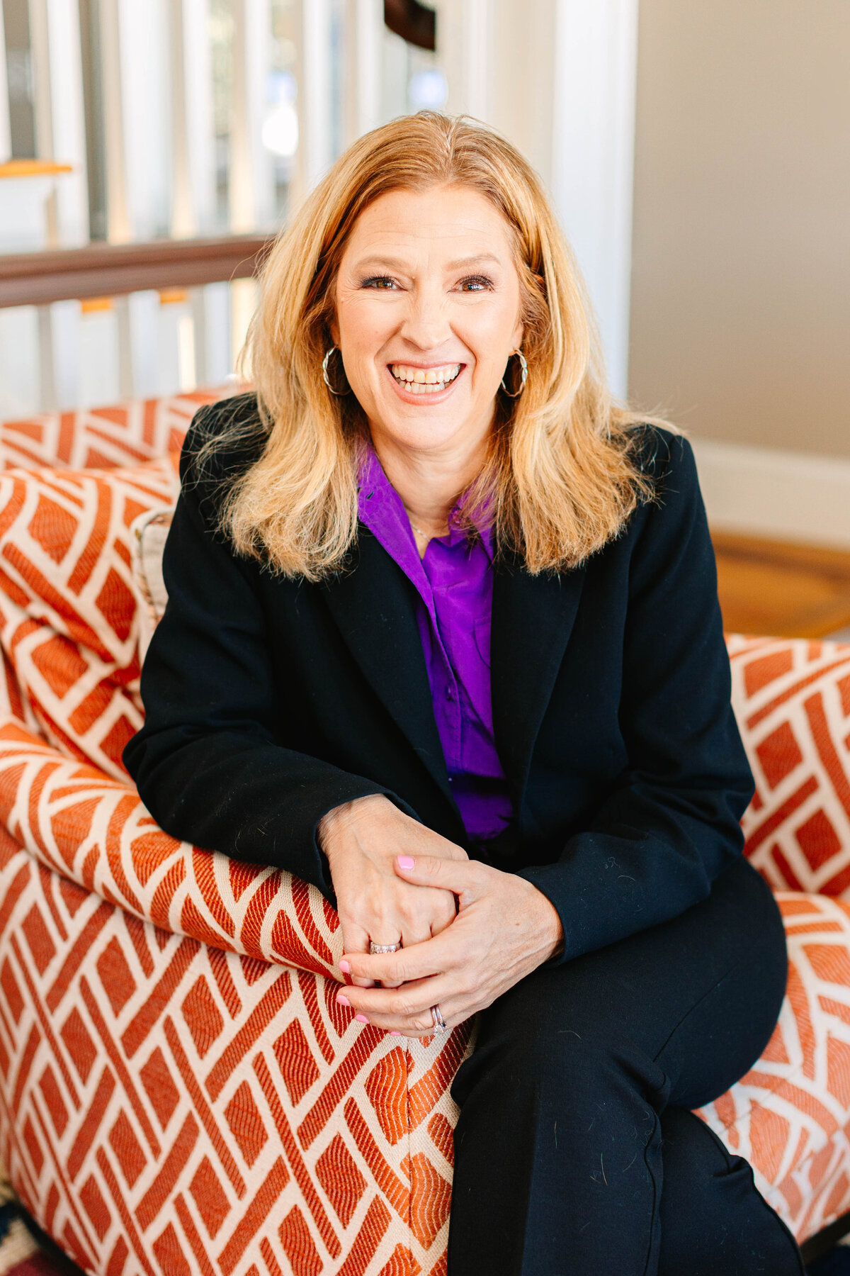 A woman with shoulder-length blonde hair sits in an orange-patterned armchair, laughing. She wears a black blazer over a vibrant purple blouse, with silver hoop earrings. The background features a warmly lit interior space.
