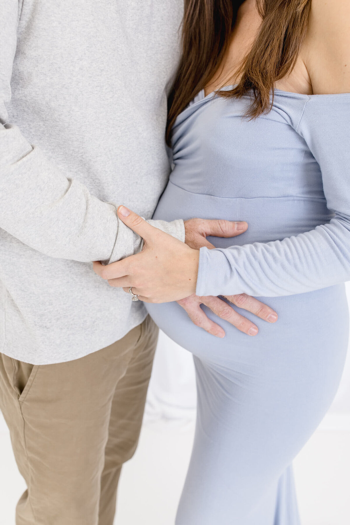 A couple stands closely, with hands gently resting on the womans pregnant belly. She is wearing a light blue off-shoulder dress, and he is wearing a light gray long-sleeve shirt and khaki pants. The background is softly lit.